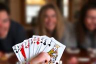 A hand holding playing cards with a friendly game in the background